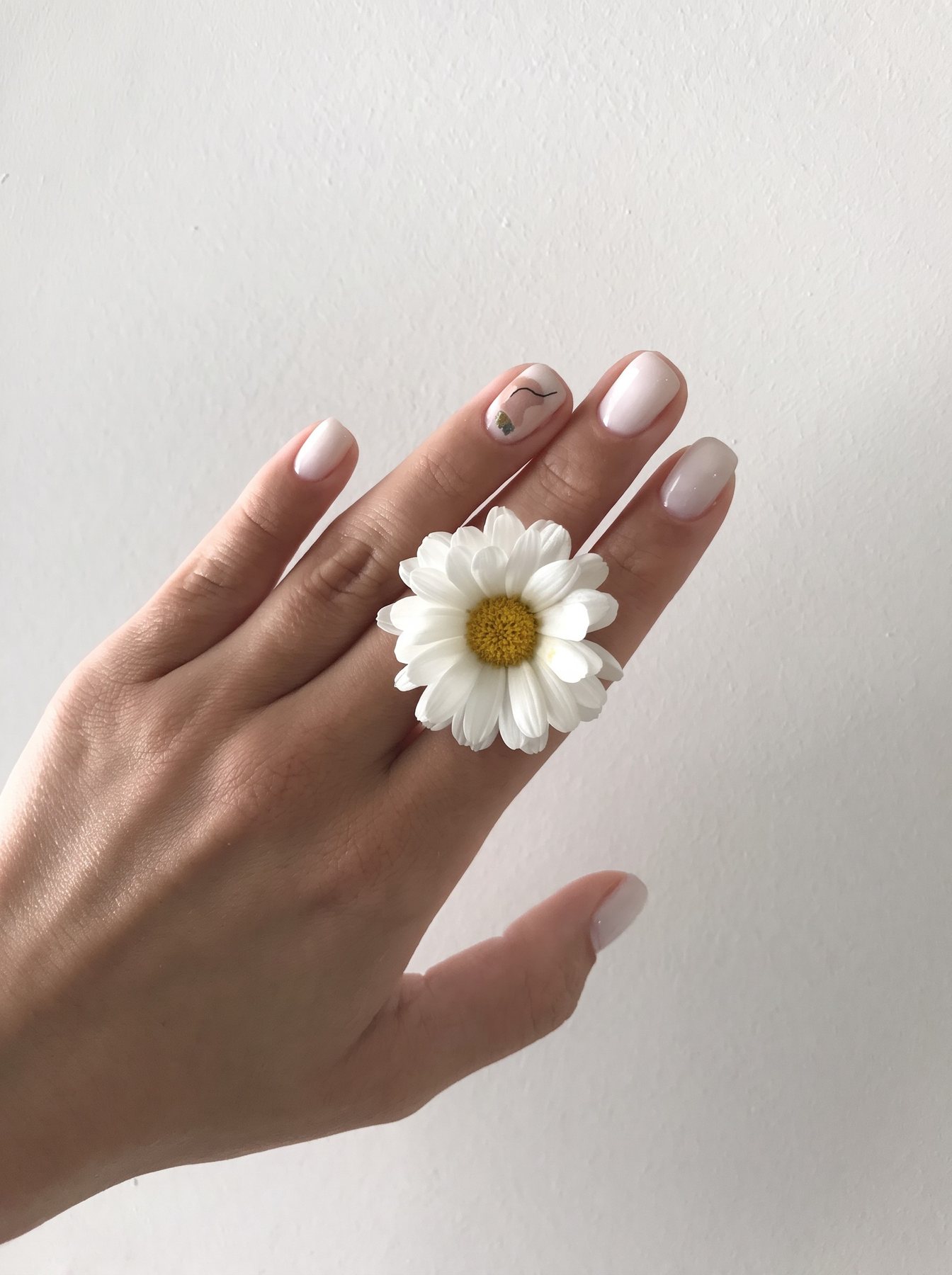 Mano con manicura en blanco lechoso y un dibujo a mano alzada en una uña, con una margarita entre los dedos